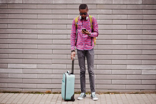 Uomo afroamericano in camicia a scacchi occhiali da sole e auricolari con valigia e zaino Viaggiatore uomo nero contro il muro che tiene telefono cellulare