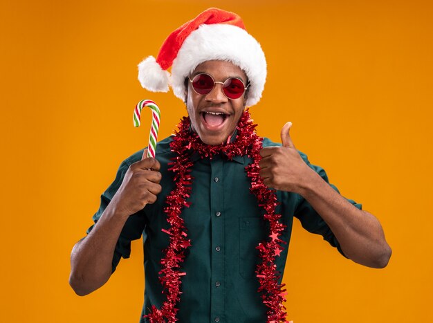 Uomo afroamericano in cappello della Santa con la ghirlanda che indossa gli occhiali che tengono il bastoncino di zucchero sorridente che mostra i pollici in su che si leva in piedi sopra la parete arancione