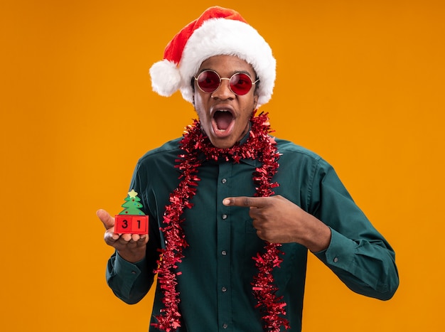 Uomo afroamericano in cappello della santa con la ghirlanda che indossa gli occhiali da sole che tengono i cubi del giocattolo con la data del nuovo anno che indica con il dito indice in piedi sorpreso e stupito sopra fondo arancio