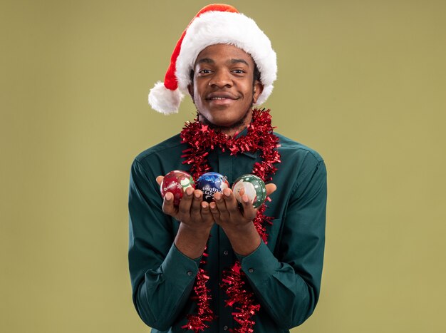 Uomo afroamericano in cappello della santa con la ghirlanda che tiene le palle di Natale che guarda l'obbiettivo sorridente con la faccia felice che sta sopra fondo verde