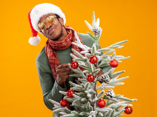 Uomo afroamericano in cappello della santa e sciarpa intorno al collo con il sorriso sul viso in piedi accanto a un albero di natale sopra la parete arancione