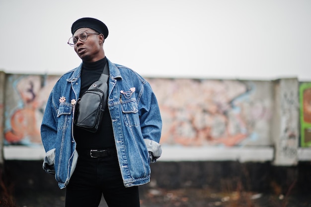 Uomo afroamericano in giacca di jeans berretto e occhiali contro il muro di graffiti sul tetto abbandonato