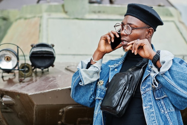 Uomo afroamericano in giacca di jeans berretto e occhiali da vista con sigaro posato contro il veicolo blindato militare btr e parlando al telefono cellulare