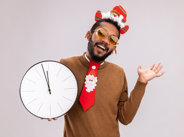 Uomo afroamericano in maglione marrone e orlo della santa sulla testa con l'orologio della tenuta della cravatta rossa divertente che guarda l'obbiettivo sorridente allegramente con il braccio alzato che sta sopra fondo bianco