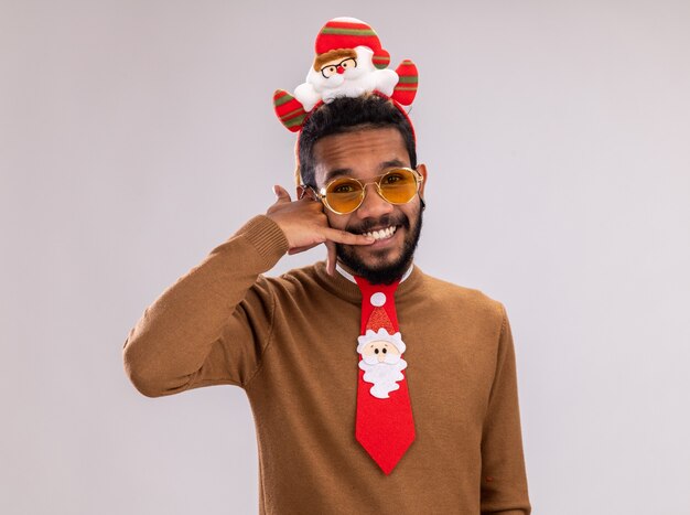 Uomo afroamericano in maglione marrone e orlo di Babbo Natale sulla testa con cravatta rossa divertente che sorride allegramente facendomi chiamare gesto in piedi su sfondo bianco