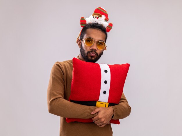 Uomo afroamericano in maglione marrone e orlo di Babbo Natale sulla testa con cravatta rossa divertente che tiene il cuscino di Natale che guarda l'obbiettivo confuso in piedi su sfondo bianco