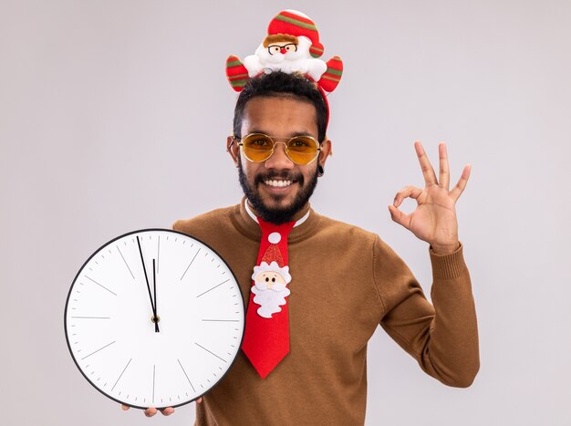 Uomo afroamericano in maglione marrone e santa orlo sulla testa con divertente cravatta rossa che tiene orologio guardando la telecamera sorridendo allegramente mostrando segno ok in piedi su sfondo bianco