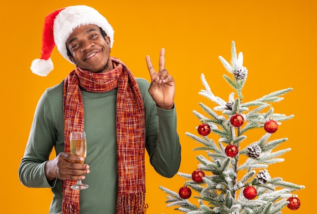 Uomo afroamericano in santa cappello e sciarpa intorno al collo tenendo un bicchiere di champagne sorridendo allegramente mostrando v-segno accanto all'albero di natale su sfondo arancione