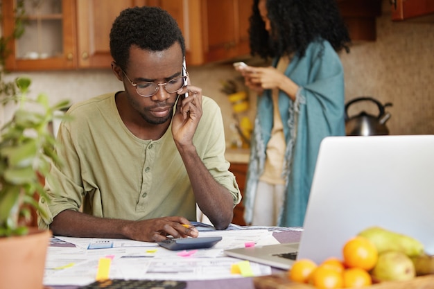 Uomo afroamericano infelice serio che ha conversazione telefonica mentre calcola il bilancio familiare in cucina