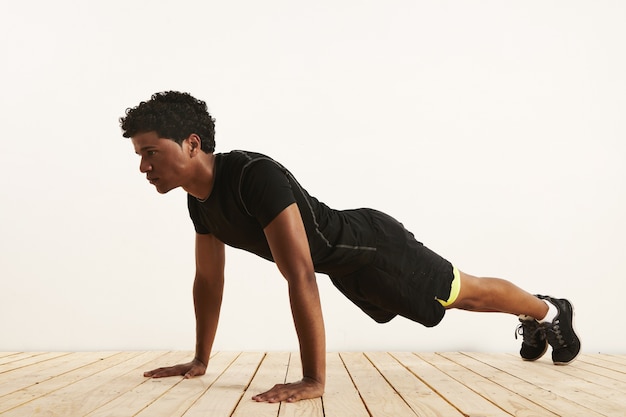 Uomo afroamericano nero in forma seria che esegue un pushup dal pavimento in legno chiaro contro un muro bianco