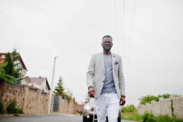 Uomo afroamericano ricco ed elegante in giacca e pantaloni bianchi occhiali posati contro l'auto suv