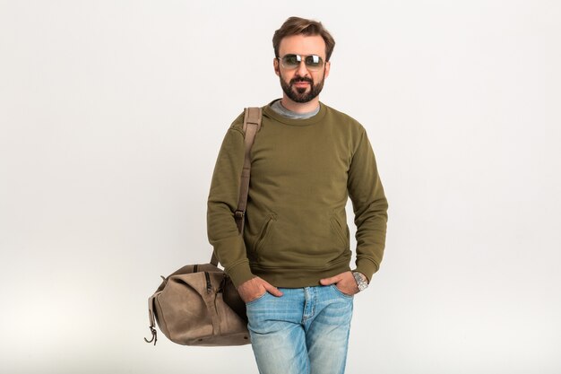 Uomo alla moda barbuto bello in posa isolato vestito di felpa con borsa da viaggio, jeans e occhiali da sole