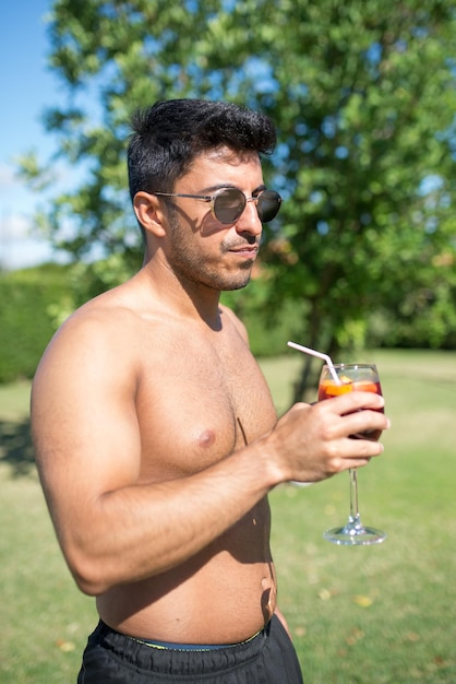 Uomo alla moda che beve cocktail luminoso. Uomo in occhiali da sole che tengono il vetro con bevanda brillante con paglia. Tempo libero, amicizia, concetto di festa