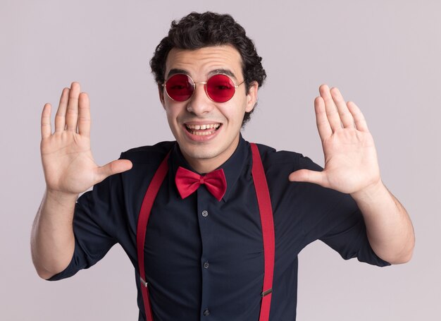 Uomo alla moda con il farfallino con gli occhiali e le bretelle guardando la parte anteriore sorridente ampiamente con le braccia alzate in piedi sul muro bianco