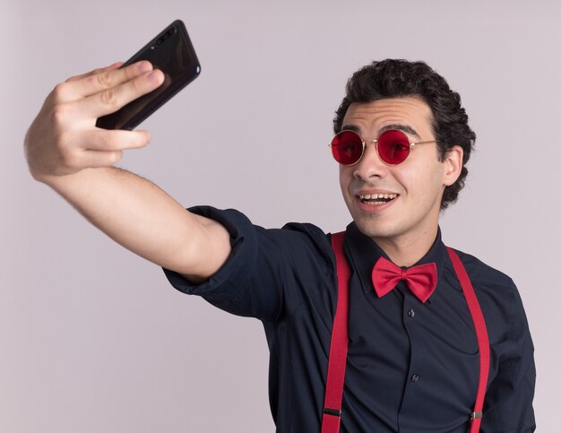 Uomo alla moda con il farfallino con gli occhiali e le bretelle utilizza lo smartphone facendo selfie sorridendo allegramente in piedi sopra il muro bianco