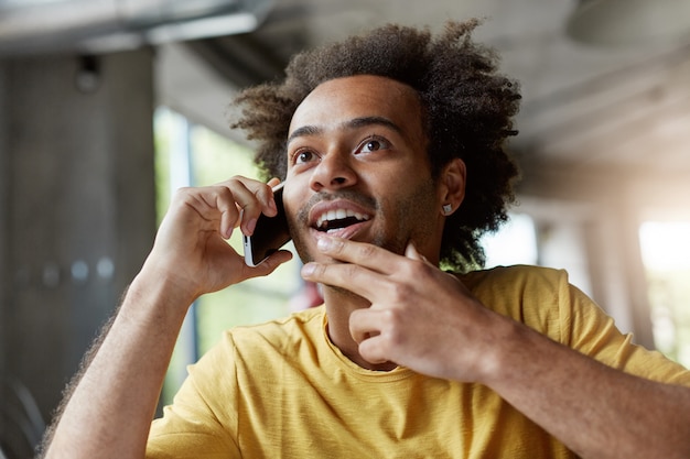 uomo alla moda con la pelle scura e capelli ricci che esamina la distanza con un aspetto misterioso mentre chiacchiera con il suo amico su smart phone.