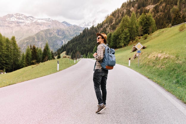 Uomo alla moda di buon umore che cammina all'aperto con lo zaino e si guarda intorno con un sorriso, godendosi il fine settimana in Italia