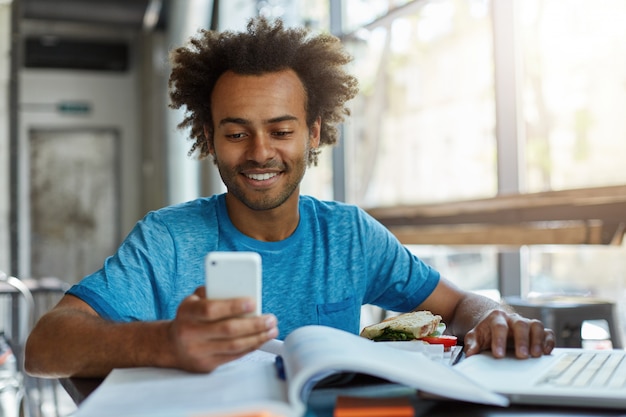 Uomo alla moda hipster con capelli scuri ricci che indossa maglietta casual blu studiando al chiuso utilizzando il telefono cellulare e computer moderni digitando messaggi al suo amico. Studente intelligente che utilizza tecnologie moderne