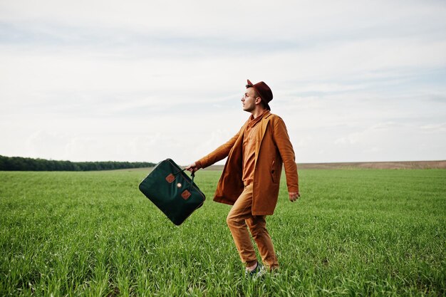 Uomo alla moda in giacca marrone occhiali e cappello con borsa poste su campo verde