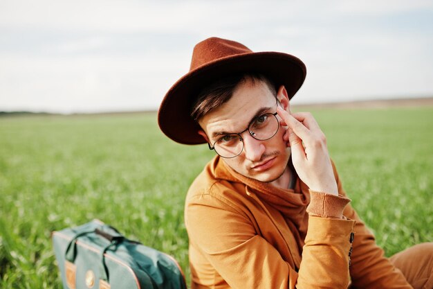 Uomo alla moda in giacca marrone occhiali e cappello con borsa poste su campo verde