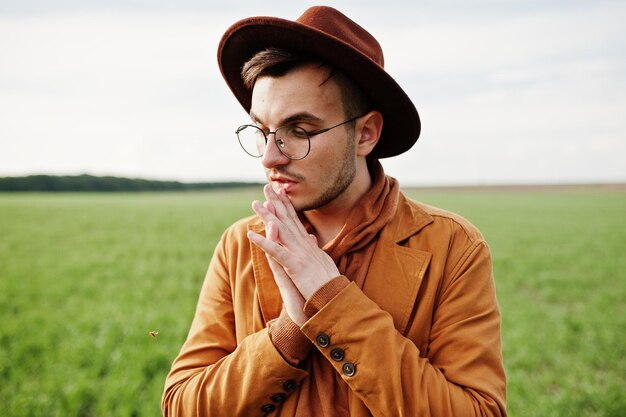 Uomo alla moda in giacca marrone occhiali e cappello posato su campo verde