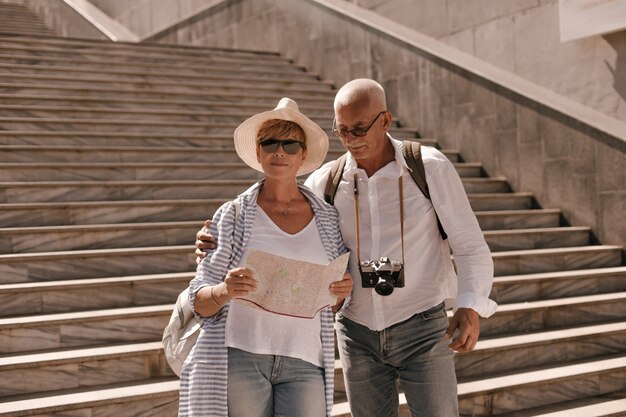 Uomo alla moda in occhiali da vista, camicia bianca e jeans con la macchina fotografica che abbraccia la moglie in cappello in camicetta a righe