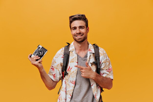 Uomo alla moda positivo in camicia leggera alla moda e maglietta grigia in posa con la macchina fotografica e sorridente su sfondo arancione isolato