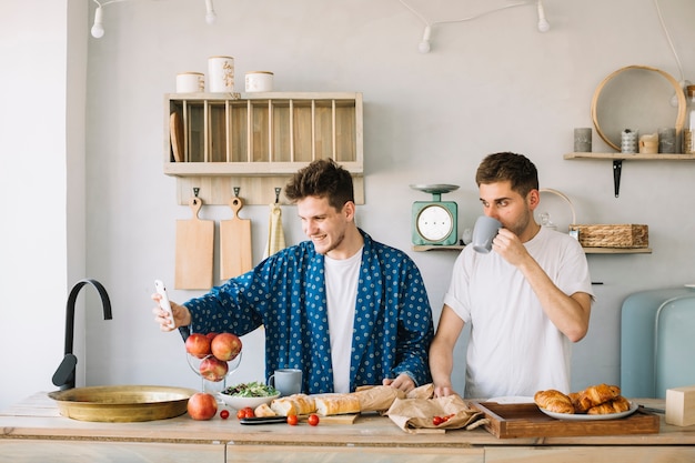 Uomo allegro che prende selfie sullo smartphone con il suo amico che beve caffè