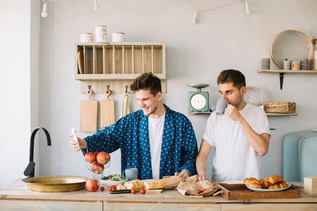 Uomo allegro che prende selfie sullo smartphone con il suo amico che beve caffè