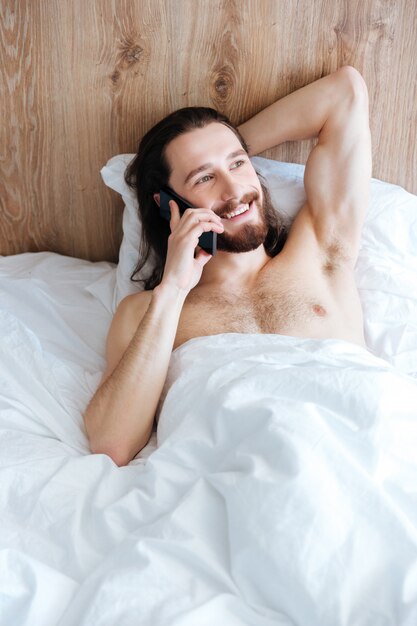 Uomo allegro che si trova a letto e che parla sul telefono cellulare