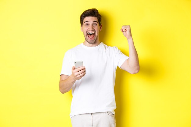 Uomo allegro che vince su smartphone, alza la mano e tiene il cellulare, raggiunge l'obiettivo dell'app, in piedi su sfondo giallo