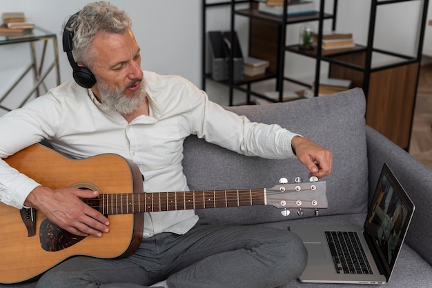 Uomo anziano a casa sul divano che usa il laptop per studiare lezioni di chitarra