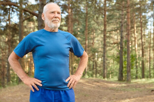 Uomo anziano autodeterminato in pensione che si allena all'aperto in legno di pino, tenendosi per mano sulla vita, facendo esercizi per riscaldare il corpo prima della corsa. Maschio barbuto in pensione riprende fiato dopo l'allenamento