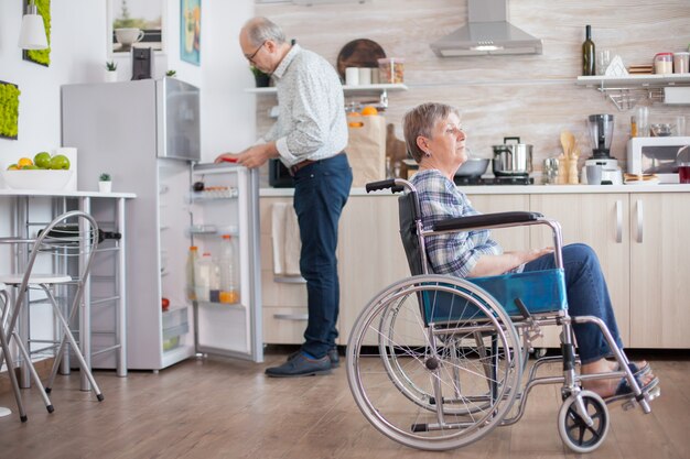 Uomo anziano che apre il frigorifero mentre sua moglie disabile è seduta su una sedia a rotelle in cucina guardando attraverso la finestra. Vivere con una persona handicappata. Marito che aiuta la moglie con disabilità. Coppia di anziani