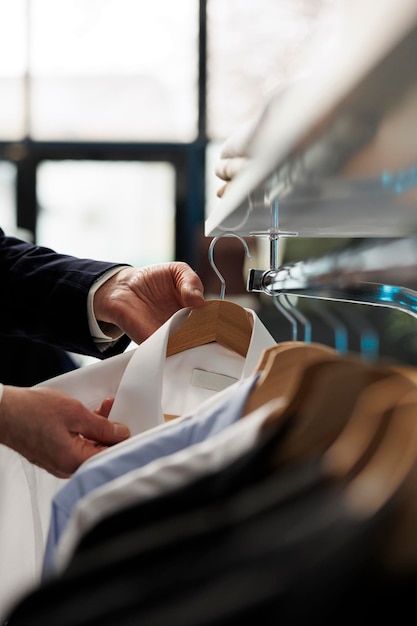 Uomo anziano che guarda grucce piene di camicie alla moda, acquistando vestiti alla moda in boutique moderne. Cliente anziano che acquista abbigliamento formale in un negozio di abbigliamento, attività commerciale. Chiuda sul colpo