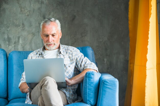 Uomo anziano che si siede sul sofà blu facendo uso del computer portatile