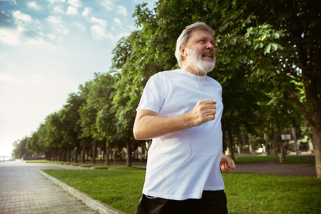 Uomo anziano come corridore con bracciale o fitness tracker in strada della città. Modello maschio caucasico che pratica corsi di jogging e cardio nella mattina d'estate. Stile di vita sano, sport, concetto di attività.