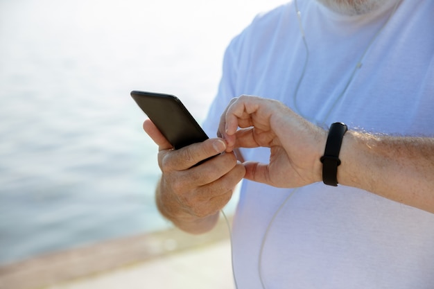 Uomo anziano come corridore con fitness tracker in riva al fiume. Modello maschio caucasico utilizzando gadget durante il jogging e l'allenamento cardio nella mattina d'estate. Stile di vita sano, sport, concetto di attività.