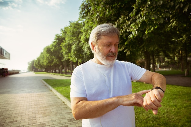 Uomo anziano come corridore con tracker di fitness in strada della città. Modello maschio caucasico utilizzando gadget durante il jogging e l'allenamento cardio nella mattina d'estate. Stile di vita sano, sport, concetto di attività.