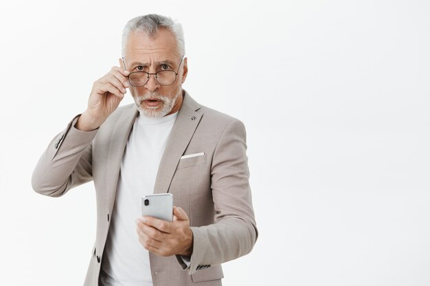 Uomo anziano confuso in vestito che tiene il telefono cellulare e alla ricerca