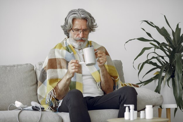 Uomo anziano da solo seduto sul divano. Uomo malato coperto di plaid. Nonno con una tazza di tè.