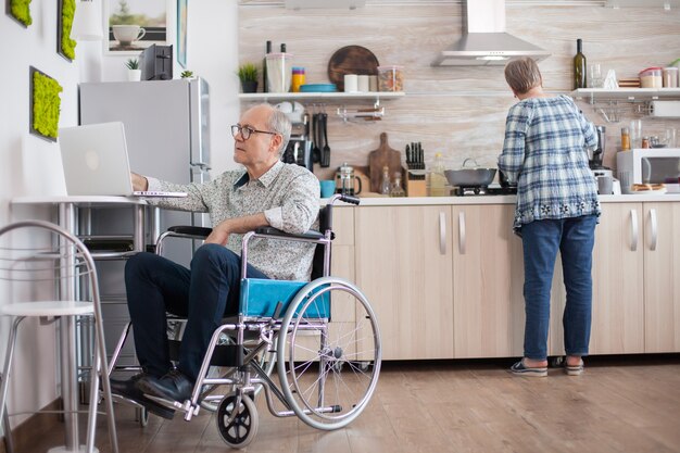 Uomo anziano disabile in sedia a rotelle che lavora al computer portatile in cucina mentre la moglie sta preparando una deliziosa colazione per entrambi. Uomo che utilizza la tecnologia moderna mentre lavora da casa.