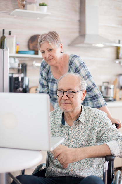 Uomo anziano disabile in sedia a rotelle e sua moglie che hanno una videoconferenza sul computer portatile in cucina. Un vecchio paralizzato e sua moglie hanno una conferenza online.