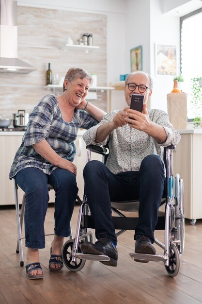 Uomo anziano disabile in sedia a rotelle e sua moglie che ridono e navigano con un moderno smartphone in cucina. Un vecchio paralizzato e sua moglie hanno una conferenza online.