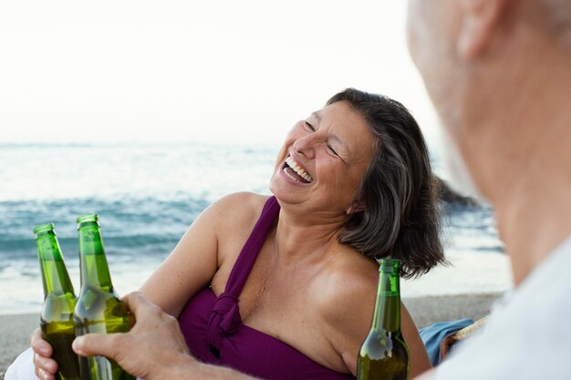 Uomo anziano e donna che ridono sulla spiaggia mentre bevono birra