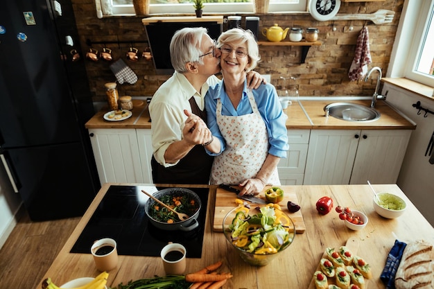 Uomo anziano felice che bacia sua moglie e si diverte mentre prepara il cibo in cucina