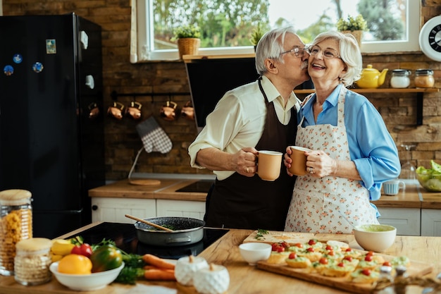 Uomo anziano felice che bacia sua moglie mentre beve caffè e prepara il cibo con lei in cucina