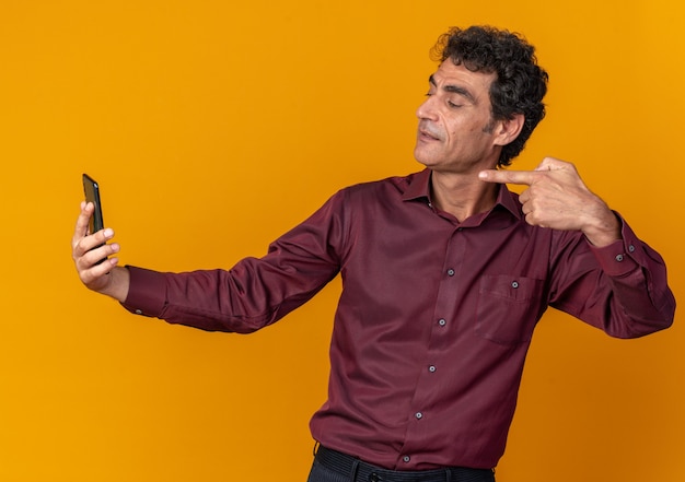 Uomo anziano in camicia viola che fa selfie utilizzando lo smartphone che sembra fiducioso puntando con il dito indice sullo schermo in piedi su sfondo arancione