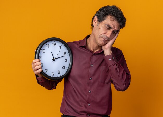 Uomo anziano in camicia viola con orologio da parete che sembra stanco e annoiato