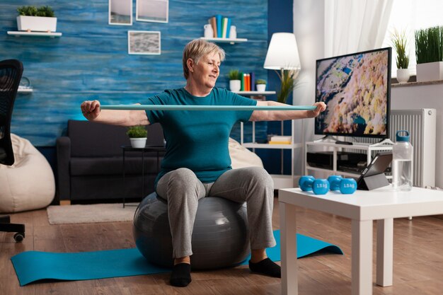 Uomo anziano in pensione seduto sulla palla svizzera fitness in soggiorno facendo allenamento fitness benessere allungando i muscoli del braccio utilizzando l'elastico di aerobica